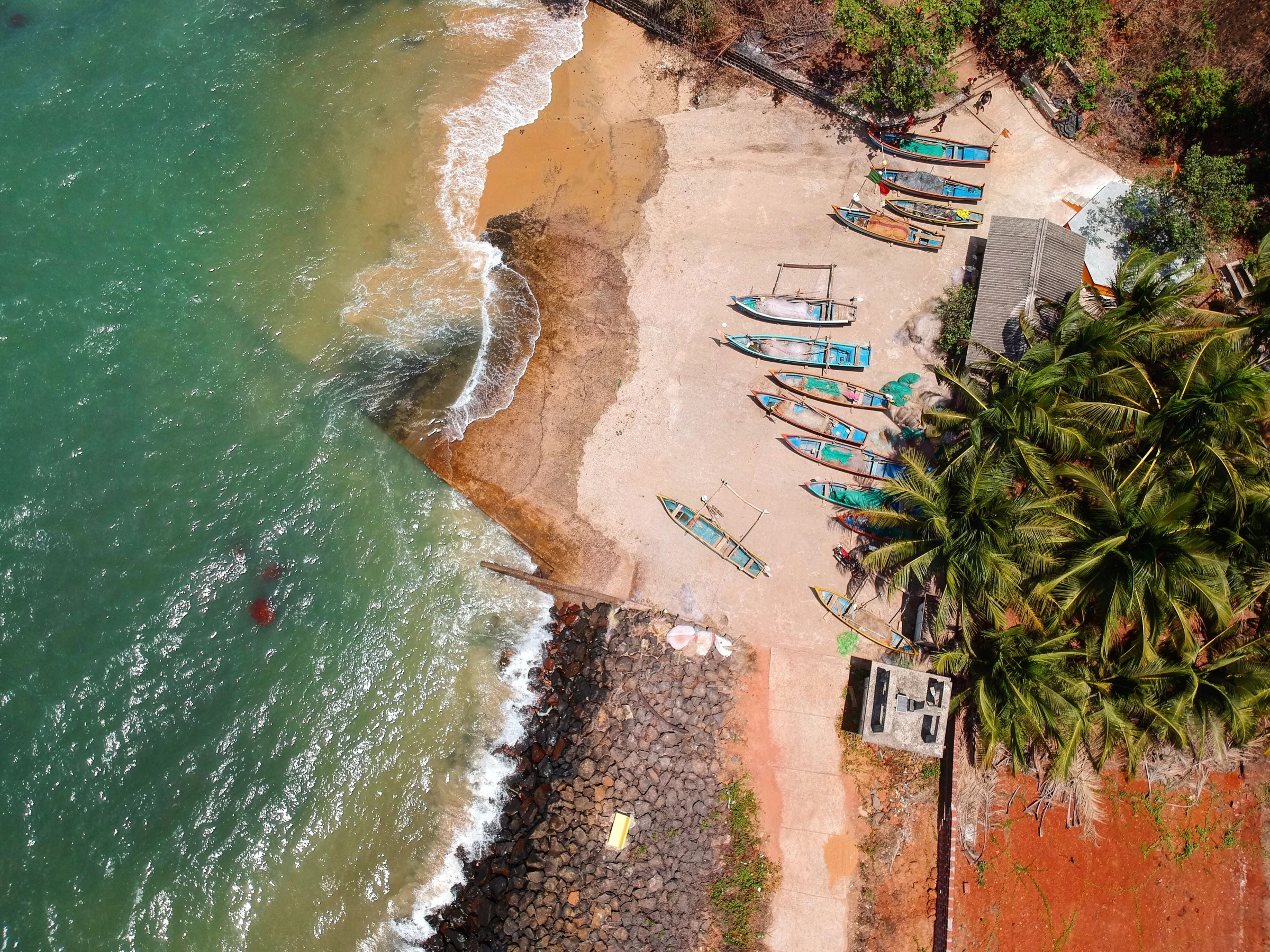 An aerial of Malvan's coast in Sindhudurg, with turquoise water, fishing boats and palms. Regional photograph, not the estate.